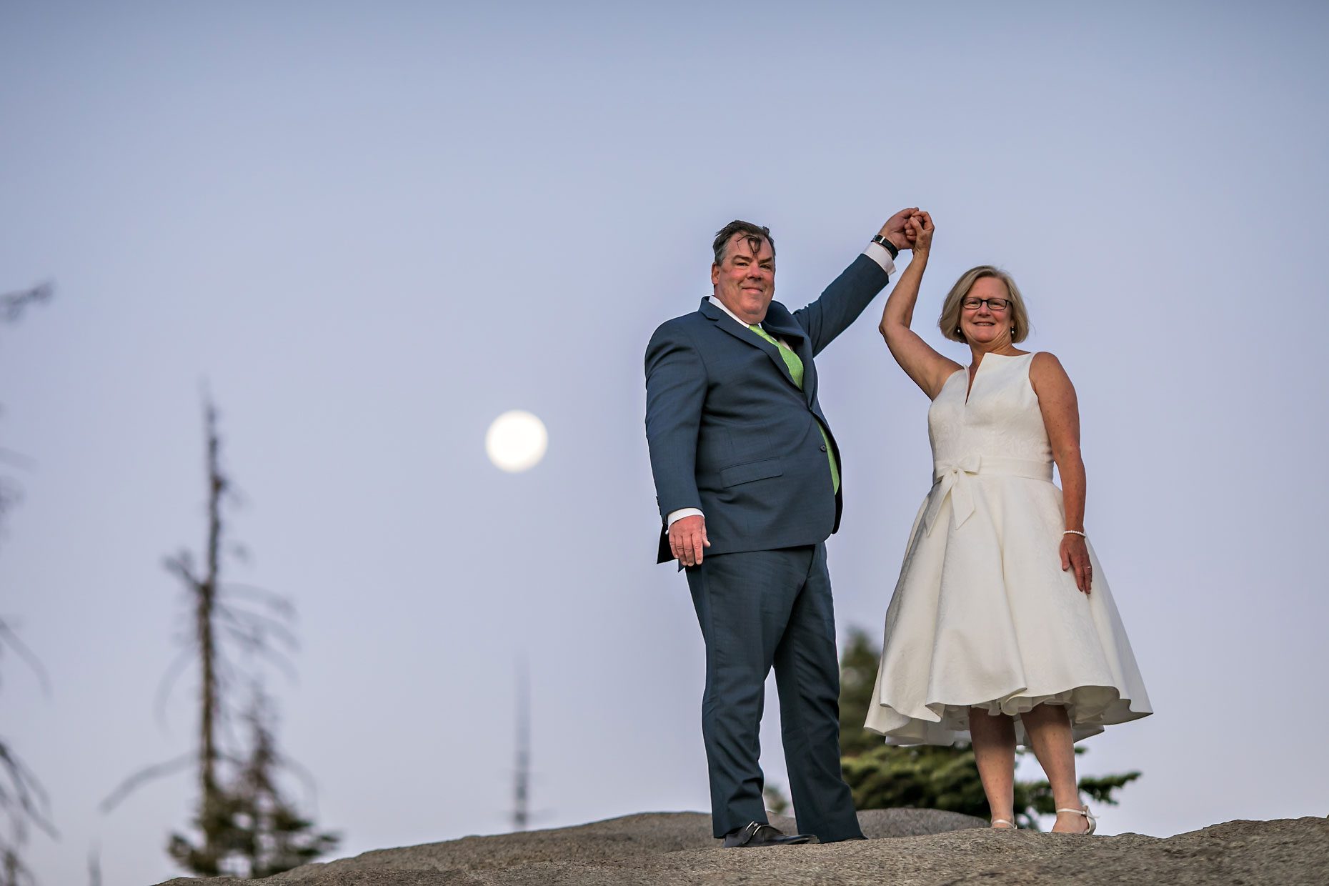 Couple gets wedding photography in Yosemite National Park at Glacier Point at sunrise with moon in background.