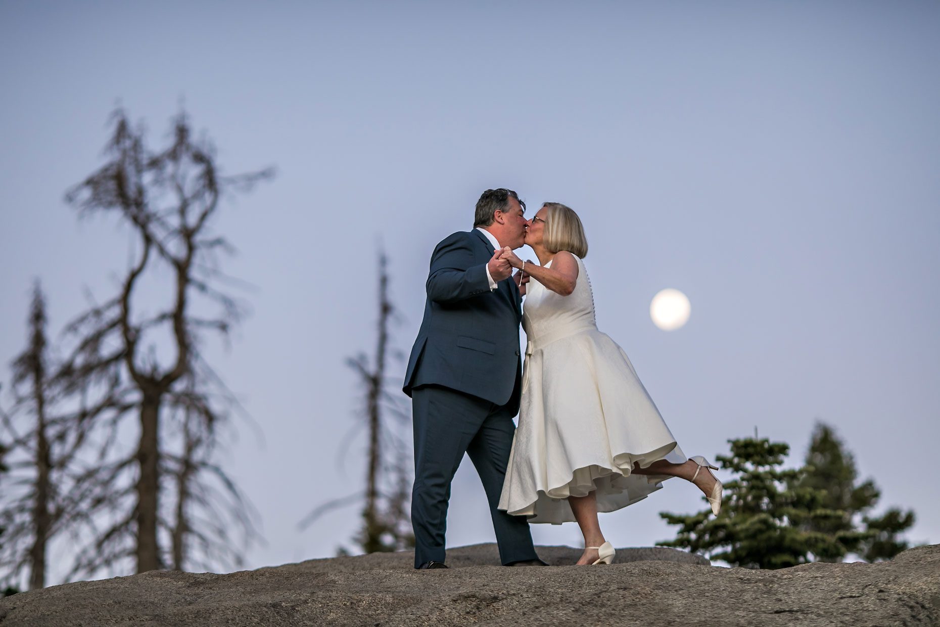 Couple gets wedding photography in Yosemite National Park at Glacier Point at sunrise with moon in background.