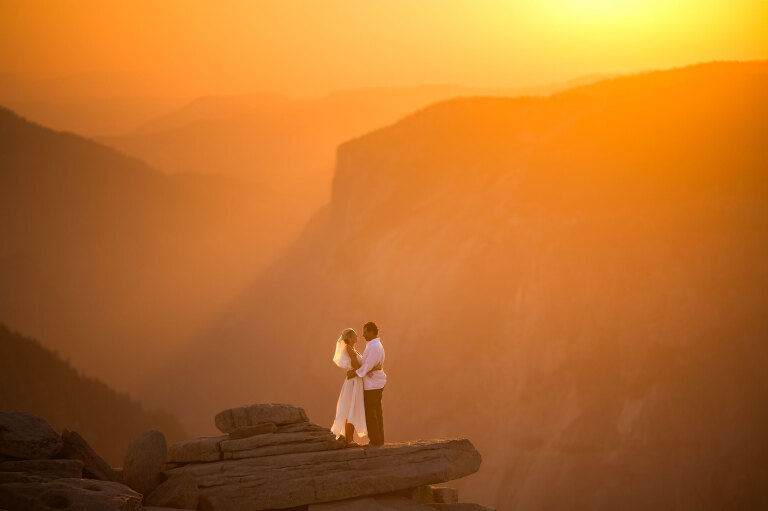 A wedding couple standing on top of a mountain kissing during sunset