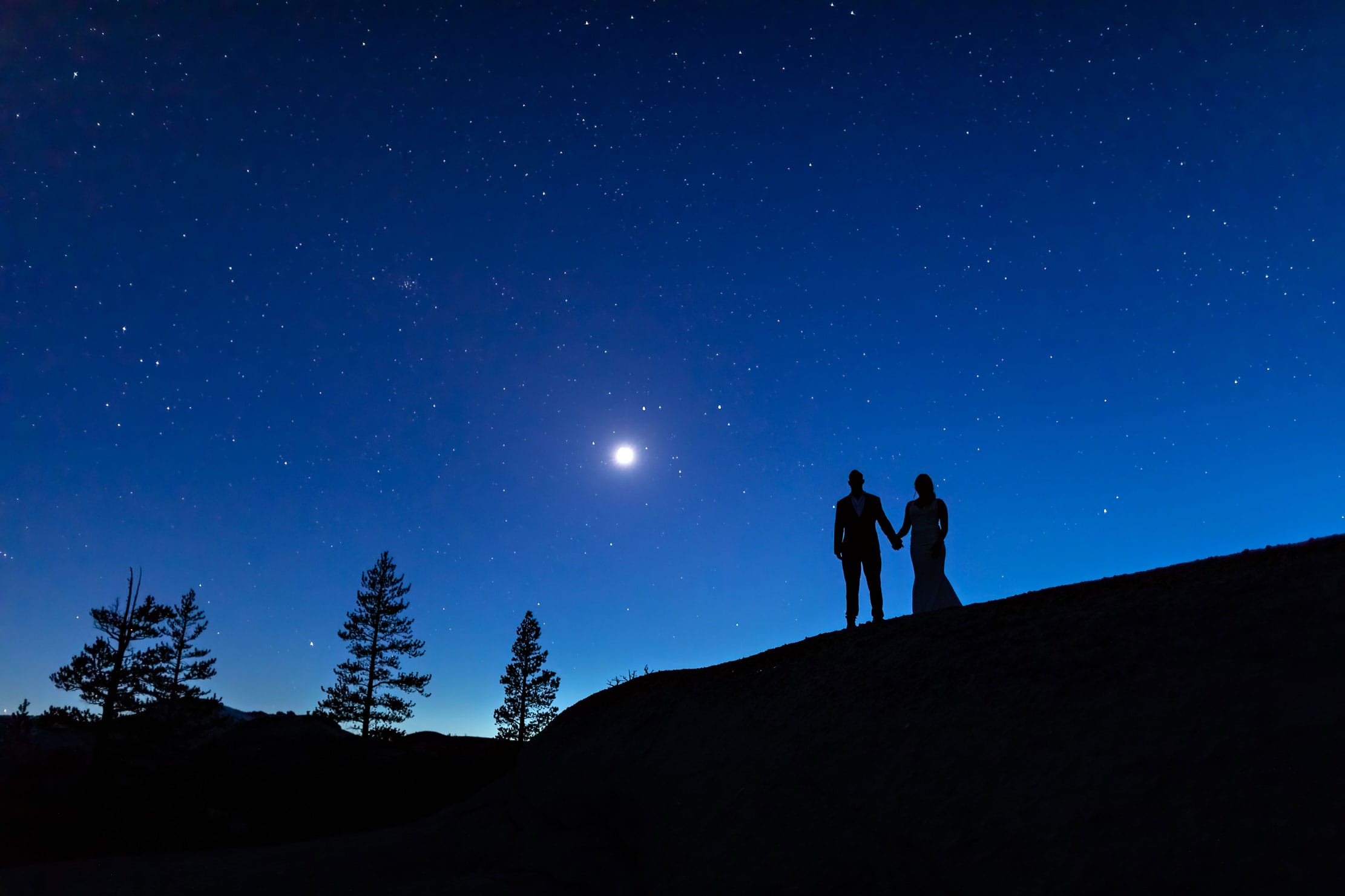 A wedding couple standing on top of mountain underneath the stars and moon.